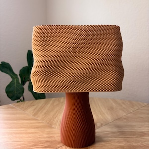 May include: A table lamp with a textured, light brown, rectangular shade featuring a wavy, undulating surface pattern. The base is a solid, darker brown, cylindrical pedestal with a fine, uniform, dotted texture covering its entire exterior.