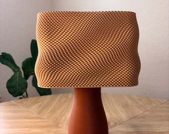 Pode incluir: Um candeeiro de mesa com um abajur retangular castanho-claro texturizado com uma superfície ondulada. A base é um pedestal cilíndrico castanho-escuro sólido com uma textura fina e uniforme de pontos em todo o seu exterior.