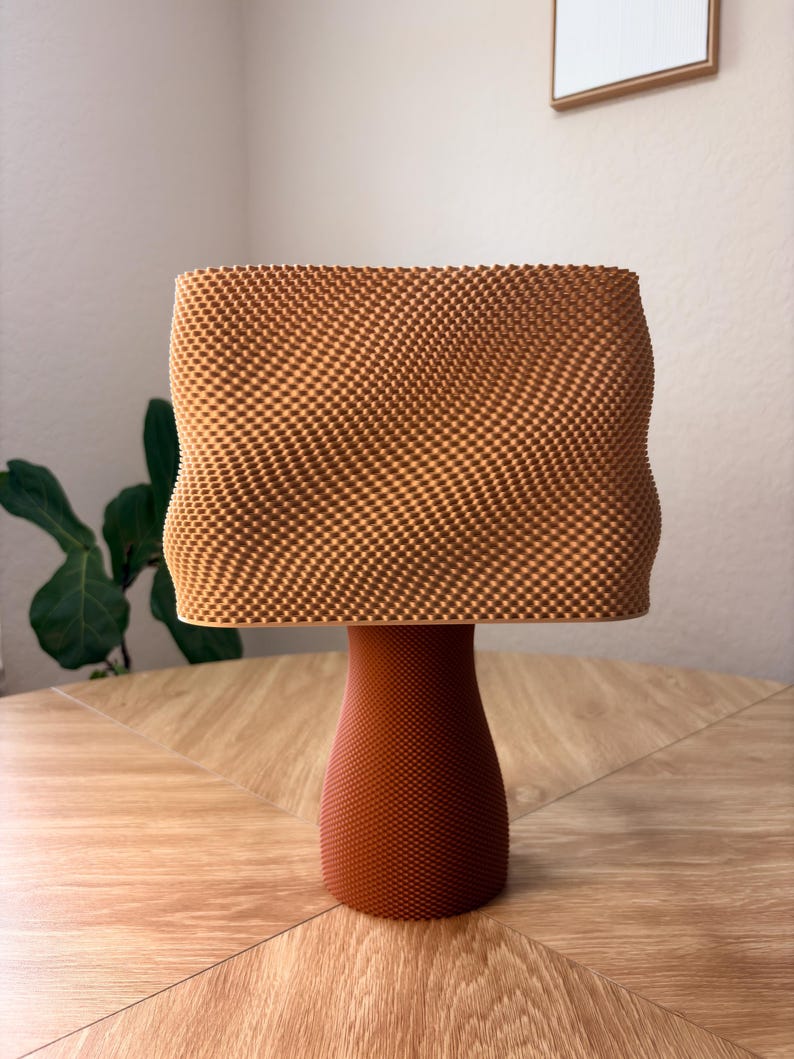 May include: A table lamp with a textured, light brown, rectangular shade featuring a wavy, undulating surface pattern. The base is a solid, darker brown, cylindrical pedestal with a fine, uniform, dotted texture covering its entire exterior.