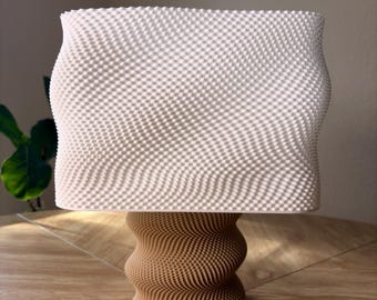 Pode incluir: Um objeto decorativo com uma secção superior retangular de cor branco-sujo com uma superfície texturada em grelha fina, suportado por uma base cilíndrica de cor castanha com um padrão geométrico ondulado e em espiral repetido.