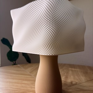 May include: A table lamp featuring a white, square-shaped shade with a textured, grid-like surface pattern. The base is a tan, cylindrical pedestal with a tapered middle and a fine, uniform dotted texture covering its entire exterior surface.