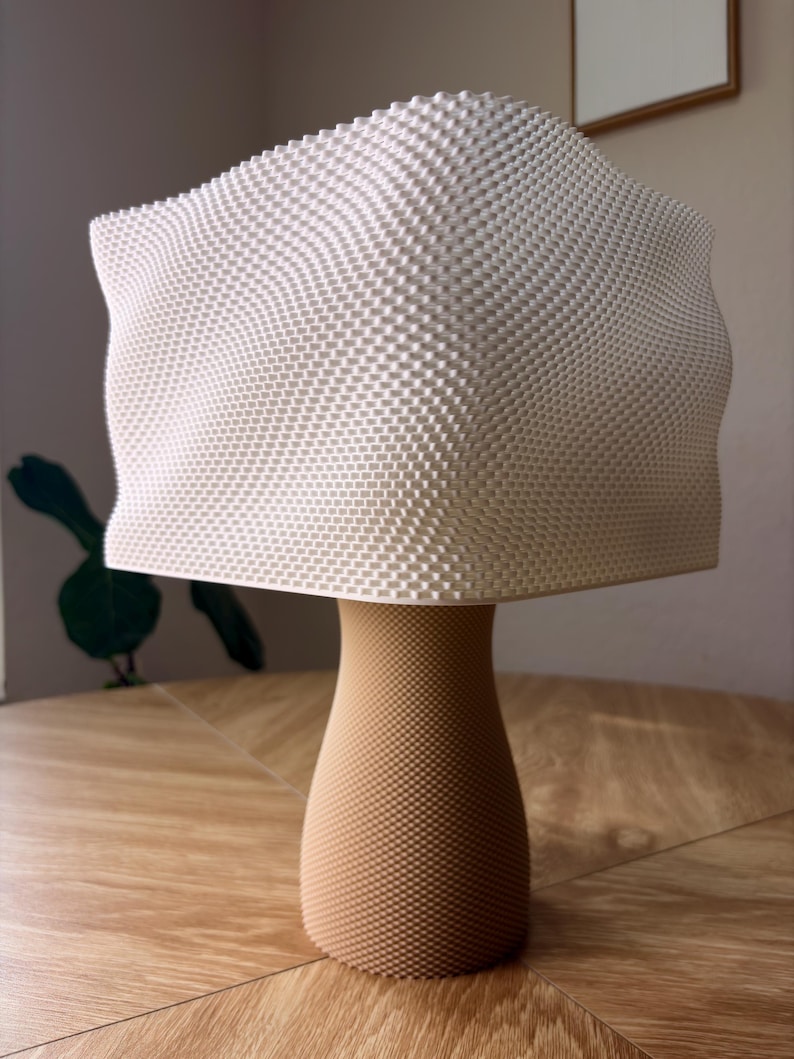 May include: A table lamp featuring a white, square-shaped shade with a textured, grid-like surface pattern. The base is a tan, cylindrical pedestal with a tapered middle and a fine, uniform dotted texture covering its entire exterior surface.
