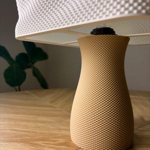 May include: A table lamp featuring a tan, hourglass-shaped base with a fine, textured grid pattern. The lamp is topped with a wide, white lampshade that also displays a consistent, raised grid texture across its entire surface area.