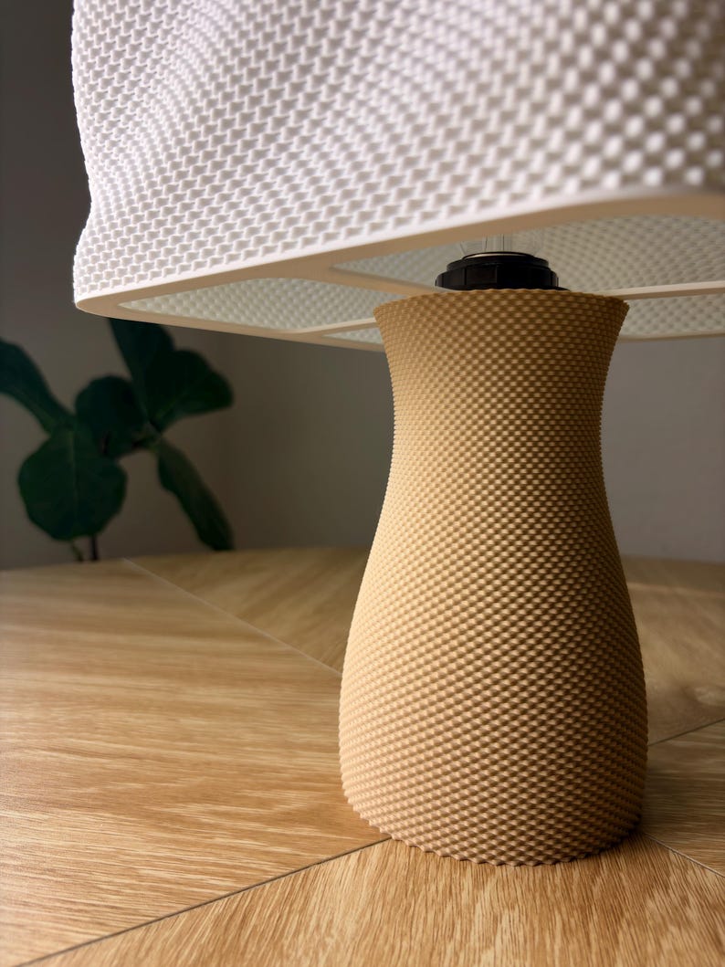May include: A table lamp featuring a tan, hourglass-shaped base with a fine, textured grid pattern. The lamp is topped with a wide, white lampshade that also displays a consistent, raised grid texture across its entire surface area.