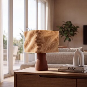 May include: A table lamp with a textured, wavy, light tan lampshade and a dark reddish-brown, tapered base with a fine dotted pattern. Next to the lamp are two stacked books labeled Design and Design Book, and a white, U-shaped ceramic vase.