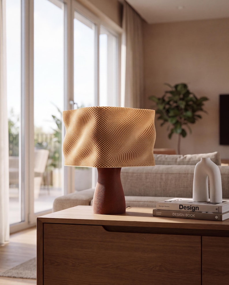 May include: A table lamp with a textured, wavy, light tan lampshade and a dark reddish-brown, tapered base with a fine dotted pattern. Next to the lamp are two stacked books labeled Design and Design Book, and a white, U-shaped ceramic vase.
