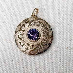 May include: A round gold-toned pendant featuring an intricate openwork filigree pattern with a textured surface. At the center of the circular design is a single, faceted, round purple gemstone set in a smooth gold-toned bezel with a small top loop.