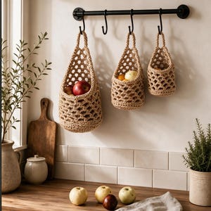 May include: Three beige, woven, hanging baskets of varying sizes suspended from a black metal wall-mounted rack with four hooks. The baskets feature an open, net-like pattern and contain various fruits, including a red apple and yellow pears.
