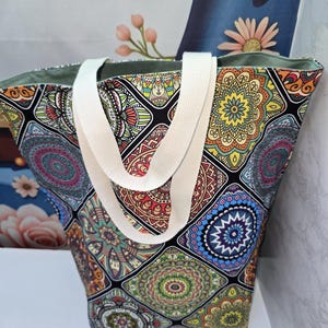 May include: A tote bag featuring a vibrant, multi-coloured mandala pattern arranged in a grid of squares. The bag has two thick, off-white woven fabric handles and a solid light green interior lining visible at the top opening of the bag.