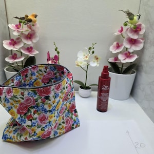 May include: A fabric pouch featuring a colourful floral pattern with pink, yellow, and blue flowers on a white background. Next to it is a dark red Wella Ultimate Repair spray bottle and a white paper with hand-drawn dimensions of 28 by 20.