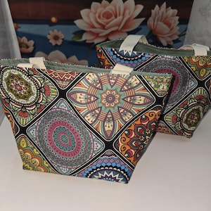 Peut inclure: Deux sacs fourre-tout arborant un motif mandala multicolore vibrant avec des formes géométriques dans des tons orange, bleu, vert et rose sur fond noir. Chaque sac possède deux anses en tissu blanc cassé et une doublure intérieure vert clair.