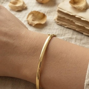 Puede incluir: Una pulsera tipo brazalete de oro pulido con una superficie lisa y redondeada, y un mecanismo de cierre de bisagra seguro. La pulsera presenta un diseño sencillo y continuo que rodea la muñeca, mostrando un acabado metálico brillante.