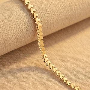 May include: A gold-toned chain bracelet composed of a continuous series of small, polished heart-shaped links connected in a linear fashion. The metallic surface reflects light, highlighting the uniform shape and smooth texture of each individual heart link.