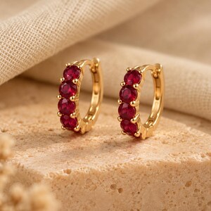 May include: A pair of gold hoop earrings, each featuring four round, deep red gemstones set in a row along the front curve. The earrings have a hinged closure mechanism and a polished metallic finish, reflecting light across the circular design.