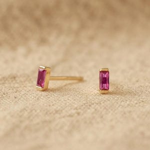 以下が含まれることがあります： 長方形のバゲットカットが施されたピンクの宝石をゴールドカラーの金属枠にはめ込んだスタッドピアス一対。それぞれのピアスには背面に細いポストが付いており、耳たぶに通して金属製のキャッチで留める構造になっています。