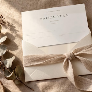 Puede incluir: Una tarjeta de regalo blanca con el texto Maison V&eacute;ra, skin, considered, colocada dentro de un sobre crema atado con una cinta de tela beige. La tarjeta presenta l&iacute;neas en blanco para cantidad, de y para, junto a una rama de eucalipto seca.