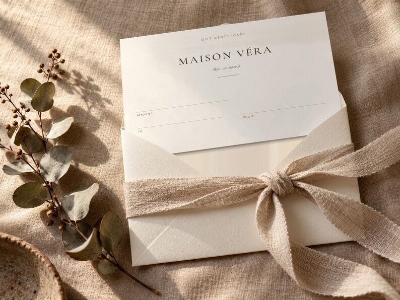 Puede incluir: Una tarjeta de regalo blanca con el texto Maison V&eacute;ra, skin, considered, colocada dentro de un sobre crema atado con una cinta de tela beige. La tarjeta presenta l&iacute;neas en blanco para cantidad, de y para, junto a una rama de eucalipto seca.