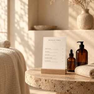 Puede incluir: Un letrero de men&uacute; rectangular de acr&iacute;lico con el texto MAISON VERA, sostenido por una base de madera clara. Junto a &eacute;l hay dos botellas de vidrio &aacute;mbar con bombas y goteros negros, sobre una mesa redonda de piedra beige texturizada.