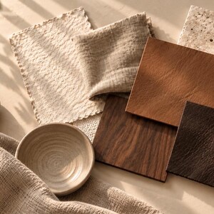 Puede incluir: Una colecci&oacute;n de muestras de materiales que incluye una tela beige texturizada, un peque&ntilde;o cuenco de cer&aacute;mica, una piedra lisa, un cuadrado de cuero marr&oacute;n claro, un cuadrado de madera veteada, un cuadrado de cuero oscuro texturizado y una baldosa de piedra beige porosa.