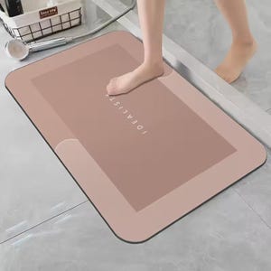 May include: A rectangular, dusty rose-colored bath mat with rounded corners and a darker central panel. The word IDEALIST is printed in white, sans-serif capital letters along the center of the mat. The mat has a smooth, flat surface texture.