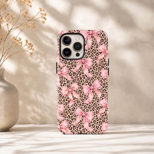 Op de afbeelding: Een rechthoekig smartphonehoesje met een herhalend patroon van lichtroze strikken tegen een bruine en zwarte luipaardprint achtergrond. Het hoesje heeft een uitsparing voor een camerasysteem met drie lenzen aan de linkerbovenkant.