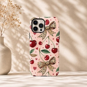 Op de afbeelding: Een telefoonhoesje met een lichtroze achtergrond met een herhalend patroon van rode kersen met groene blaadjes, strikjes met luipaardprint en kleine rode hartjes. Het hoesje heeft een zwarte camera-uitsparing en een matte afwerking.