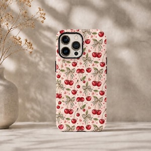 Op de afbeelding: Een smartphonehoesje met een herhalend patroon van felrode kersen gecombineerd met strikjes met luipaardprint op een lichtroze achtergrond, verspreid met kleine rode sterretjes en hartjes. Het hoesje heeft een zwarte camera-uitsparing.