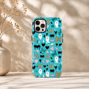 Op de afbeelding: Een turquoise telefoonhoesje met een herhalend patroon van witte spoken, strikjes met luipaardprint, zwarte harten, roze harten en zwarte verfdruppelvormen. Het hoesje heeft een rechthoekige uitsparing voor de camera met drie lenzen.