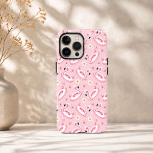 May include: A pink smartphone case featuring a repeating pattern of white and pink flamingo-shaped pool floats interspersed with small white and yellow tropical flowers. The case has a black camera lens cutout on the upper left corner.