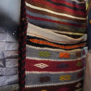 Puede incluir: Un tapiz tejido con rayas horizontales en tonos rojo, marrón, gris y blanco. El textil incluye un patrón repetitivo de pequeños rombos en amarillo, verde y naranja, con un borde trenzado en negro y rojo a lo largo del lateral.