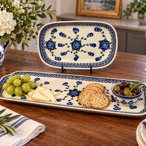 May include: A rectangular ceramic serving platter and an oval ceramic platter, both featuring a cream-colored base with a blue floral pattern and a decorative blue border. The rectangular platter holds green grapes, cheese slices, crackers, and a small bowl.