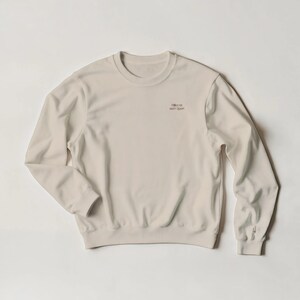 May include: A cream-colored crewneck sweatshirt featuring long sleeves and a ribbed collar, cuffs, and hem. The chest area displays the small, dark-colored text "Stillen ist auch Sport" printed on the upper right side of the garment.