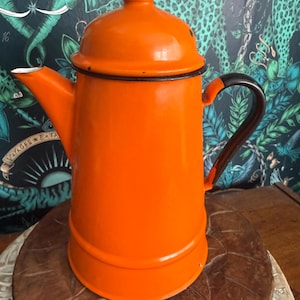 May include: A bright orange enamel coffee pot with a matching lid and a dark brown handle. The pot features a tapered cylindrical body, a wide base, and a pointed spout. The rim of the lid and the base of the handle are accented with thin black trim.