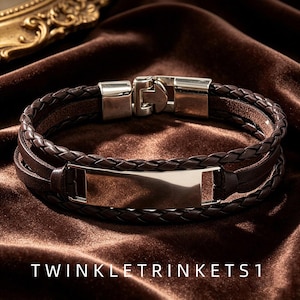 Könnte beinhalten: Ein mehrsträngiges Armband mit dunkelbraunen geflochtenen Lederbändern und einer glatten, polierten rechteckigen ID-Platte aus silberfarbenem Metall. Das Armband wird mit einem passenden silberfarbenen Metallverschluss geschlossen. Der Text TWINKLETRINKETS1 ist sichtbar.