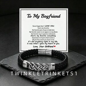 Könnte beinhalten: Ein schwarzes geflochtenes Lederarmband mit einem silberfarbenen Metallverschluss in komplexem Knotendesign. Der Verschluss ist mit LOVE YOU und Y.B. graviert. Eine weiße Karte zeigt den Text To My Boyfriend und eine Liebesbotschaft.
