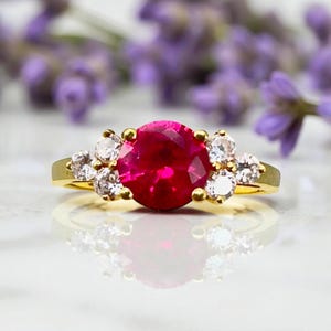 May include: A gold ring featuring a large, round, vibrant red gemstone as the centerpiece, flanked by smaller, clear gemstones. The ring's band is a polished gold, and the setting highlights the brilliance of the stones. The ring is set against a blurred background of purple flowers.