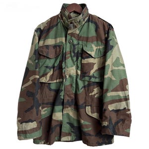 May include: A camouflage field jacket featuring a woodland pattern in shades of green, brown, and black. The jacket includes a high collar with a zipper, a front button closure, two chest pockets with flaps, and two lower pockets.
