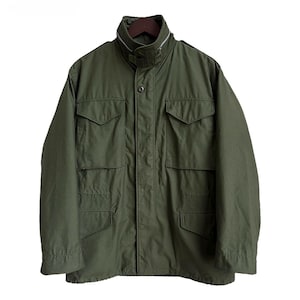 May include: An olive green field jacket featuring a high collar with a visible zip, a central button placket, and four large front pockets with buttoned flaps. The garment has long sleeves and a structured, utilitarian design.