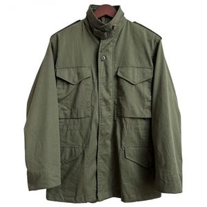 May include: An olive green field jacket featuring a high collar with a zipper, a front button closure, and four large rectangular pockets with buttoned flaps on the chest and waist. The jacket includes shoulder epaulets and long sleeves.