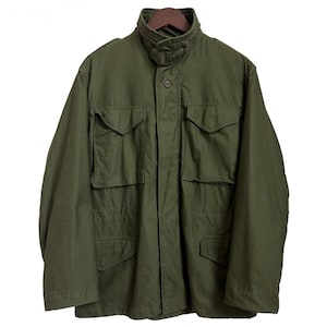 May include: An olive green field jacket featuring a high stand-up collar with a tab closure, a front zip concealed by a buttoned placket, and four large rectangular flap pockets on the front. The jacket has long sleeves and a structured fit.