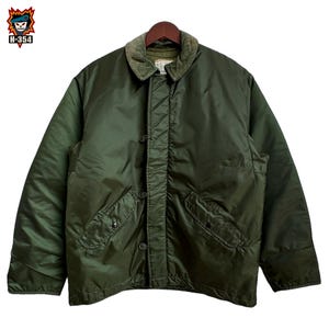 May include: An olive green jacket featuring a corduroy collar, a front button closure with a storm flap, and two slanted side pockets with buttoned flaps. The jacket has long sleeves and a smooth, quilted-style finish on the outer shell.