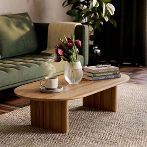 Op de afbeelding: Een ovale houten salontafel met twee dikke, gebogen poten. Op de tafel staat een dampende keramische mok op een schoteltje, een doorzichtige glazen vaas met donkerrode tulpen en een stapel van drie boeken met daarop een leesbril.