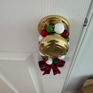 May include: A gold-colored metal doorknob adorned with a circular string of wooden beads in red, white, green, and red-and-black buffalo plaid patterns, finished with a matching red-and-black buffalo plaid fabric bow hanging at the bottom.