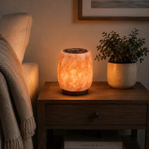 May include: A cylindrical salt lamp with a rough, translucent orange texture and a dark circular base. The top of the lamp features a metal rim containing small, dark, textured pieces. The lamp emits a warm, glowing light from within.