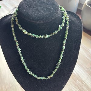 Könnte beinhalten: Eine lange Halskette, die aus zahlreichen kleinen, unregelmäßigen, getrommelten grünen Steinfragmenten besteht. Die Kette ist zweifach um das Display gelegt und zeigt die verschiedenen Schattierungen von Wald- und Smaragdgrün der Rohsteine.