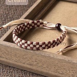 May include: A woven bracelet featuring a repeating checkered pattern in brown and cream thread. The bracelet includes adjustable tie strings and a single dark brown bead. The text "Brown" and "Cream" is visible in the upper left corner.
