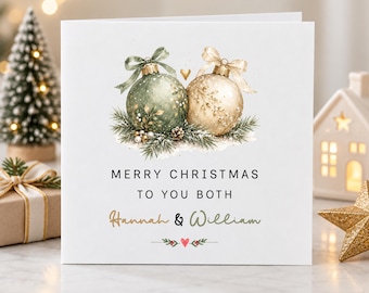 May include: A square white Christmas greeting card featuring two illustrated ornaments, one sage green and one cream, tied with ribbons and resting on pine needles. The text reads Merry Christmas to you both, Hannah & William.