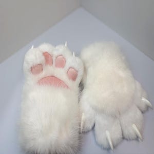 May include: A pair of white faux fur gloves designed to resemble animal paws. The gloves feature soft, plush white material with light pink padded paw pads on the palm side and pointed, white claw-like accents extending from the fingertips.