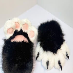 May include: A pair of plush animal paw gloves featuring long black and white faux fur. One glove displays a palm with soft pink pads, while the other glove shows white fur-covered fingers tipped with sharp, curved black plastic claws.