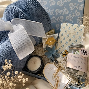 May include: A gift box containing a blue crocheted blanket with a white ribbon, a 2026-2027 floral planner, a jar of Summer Berry tea, a Crisp White candle, a blue flower hair clip, two gold hair clips on a card, and a gold lip-shaped item.